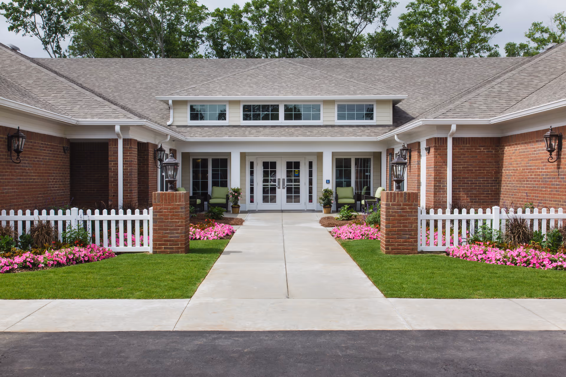 Front entrance of a senior living facility with a concrete walkway leading to double glass doors. The building has a brick exterior with white trim, a white picket fence, green lawn, and pink flowers on either side of the walkway. There are outdoor chairs and potted plants near the entrance.