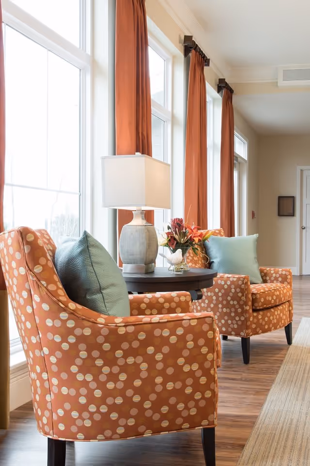 Two orange armchairs with a polka dot pattern and light blue pillows are positioned next to a round wooden table with a lamp and a floral arrangement. The seating area is by tall windows with long rust-colored curtains, and the floor is wooden with a beige rug.