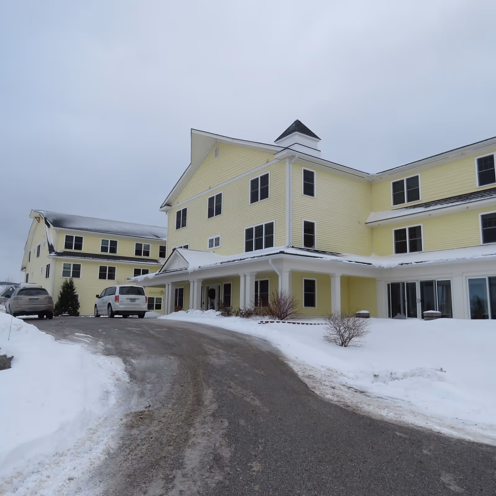 Yellow senior living building with snow-covered driveway and parked cars at entrance.