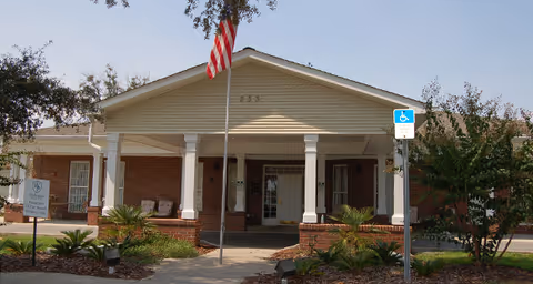 Front entrance of a single-story brick senior living facility with a covered portico, columns, an American flag, and landscaping.