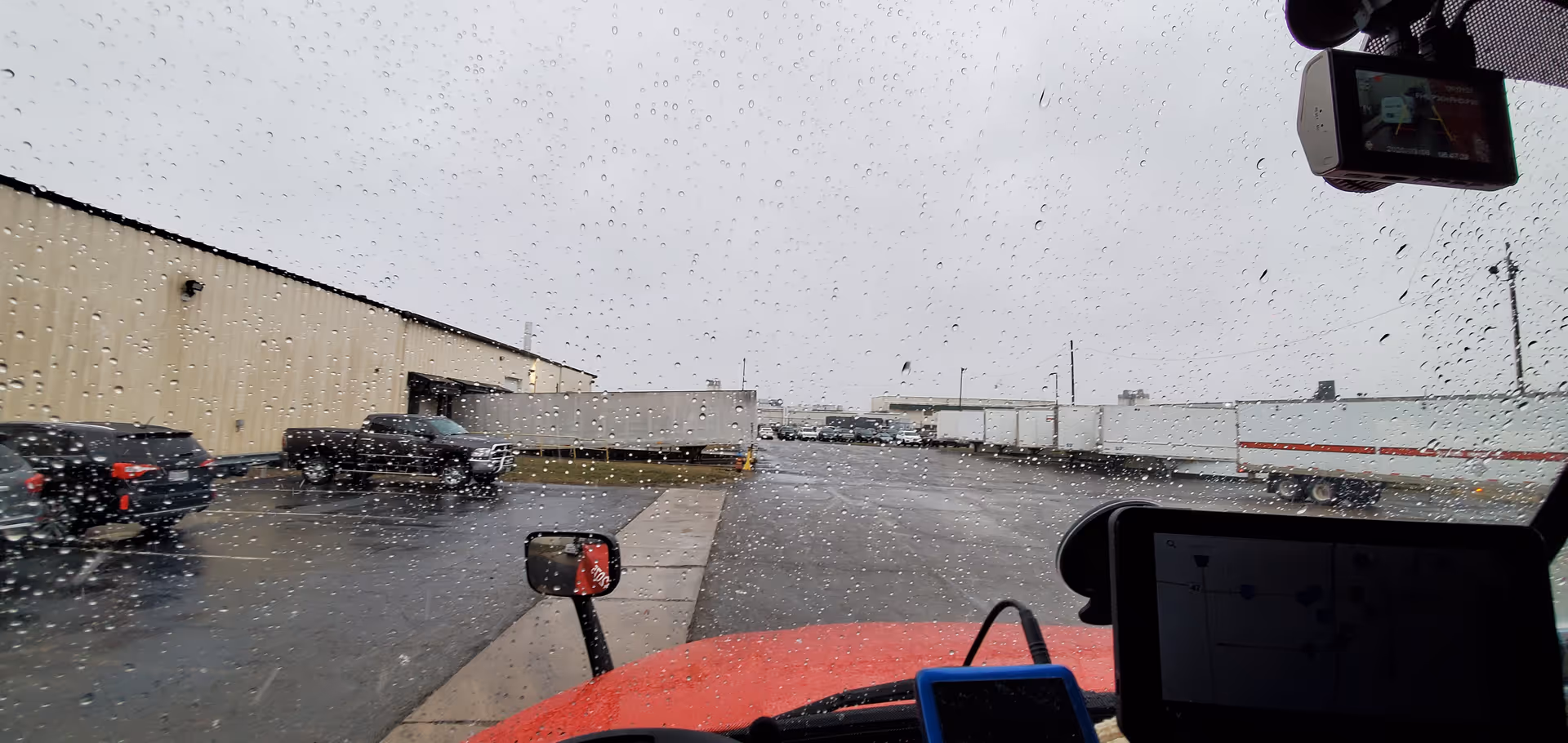 Rainy view from inside a vehicle showing a parking lot and industrial buildings.