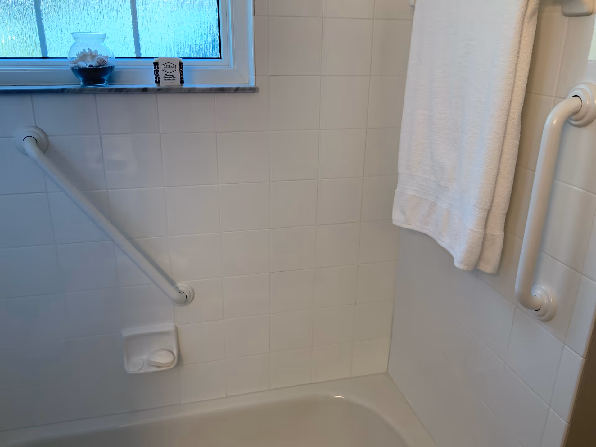 A white-tiled bathroom featuring safety grab bars, a towel, and a small fish bowl by window.