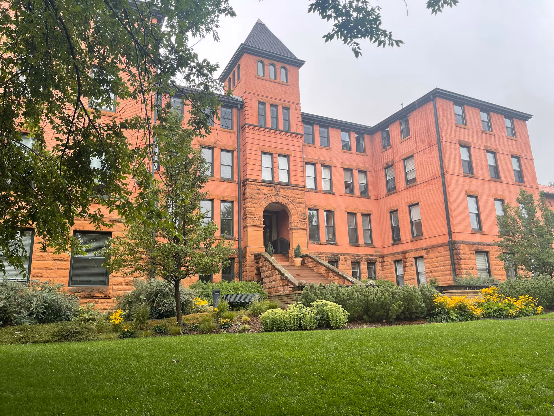 Red brick senior living building with arched entrance, steps, and manicured lawn.