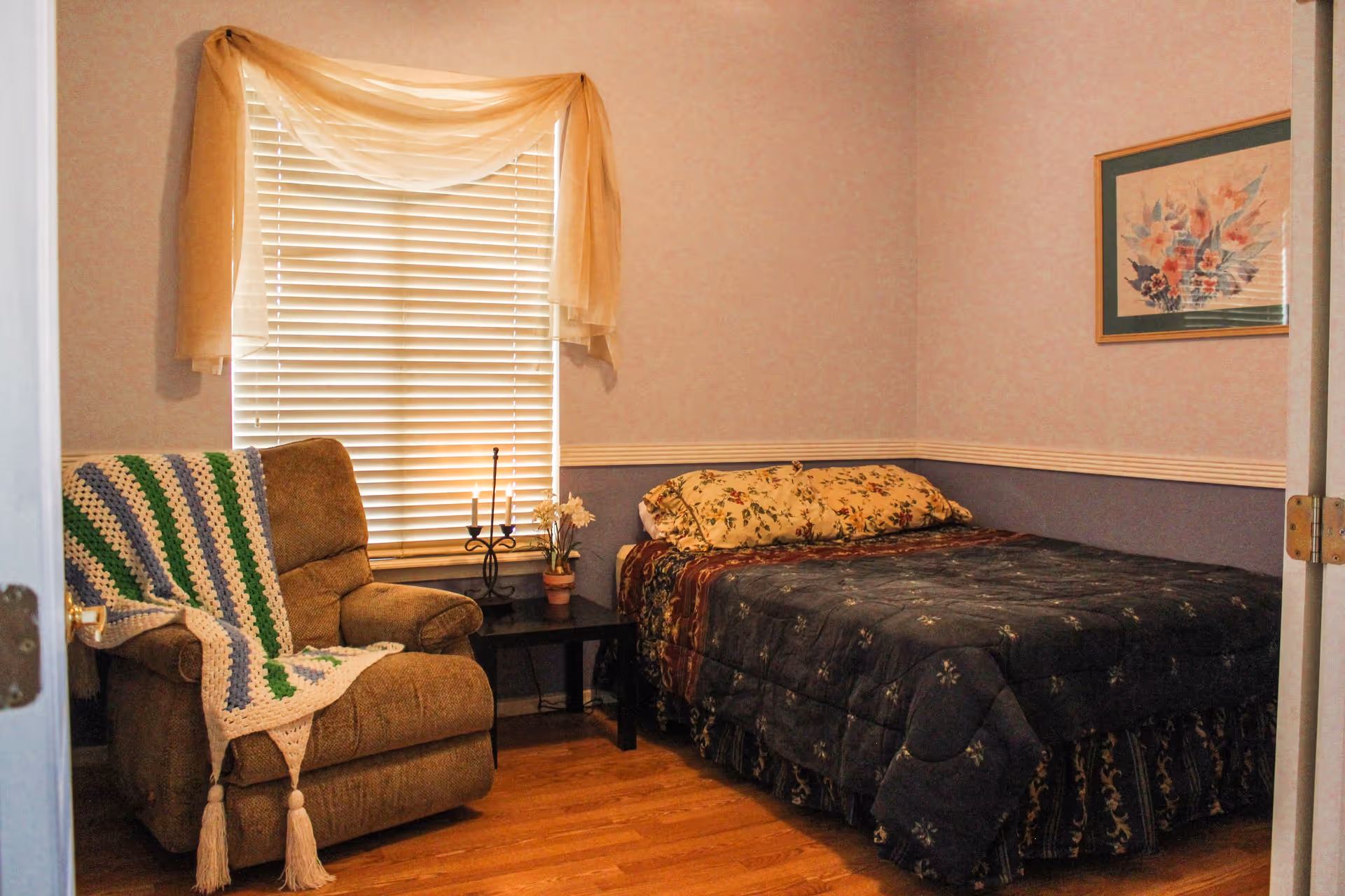 A cozy bedroom with a bed covered in a dark floral comforter and matching pillows. Next to the bed is a small black side table with a potted plant and a candle holder with two candles. A comfortable brown armchair with a crocheted blanket draped over it is positioned near a window with closed blinds and a sheer curtain. A framed floral painting hangs on the wall above the bed. The room has wooden flooring and soft pastel-colored walls.
