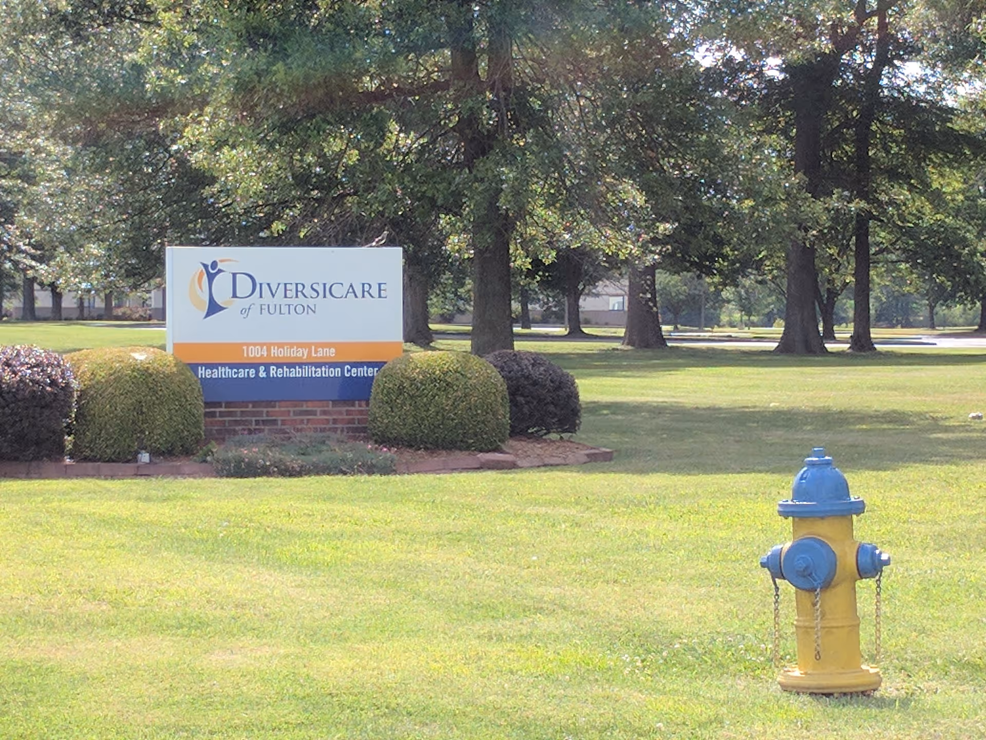 Outdoor lawn scene with Diversicare of Fulton sign, trimmed bushes, trees, and a yellow fire hydrant.