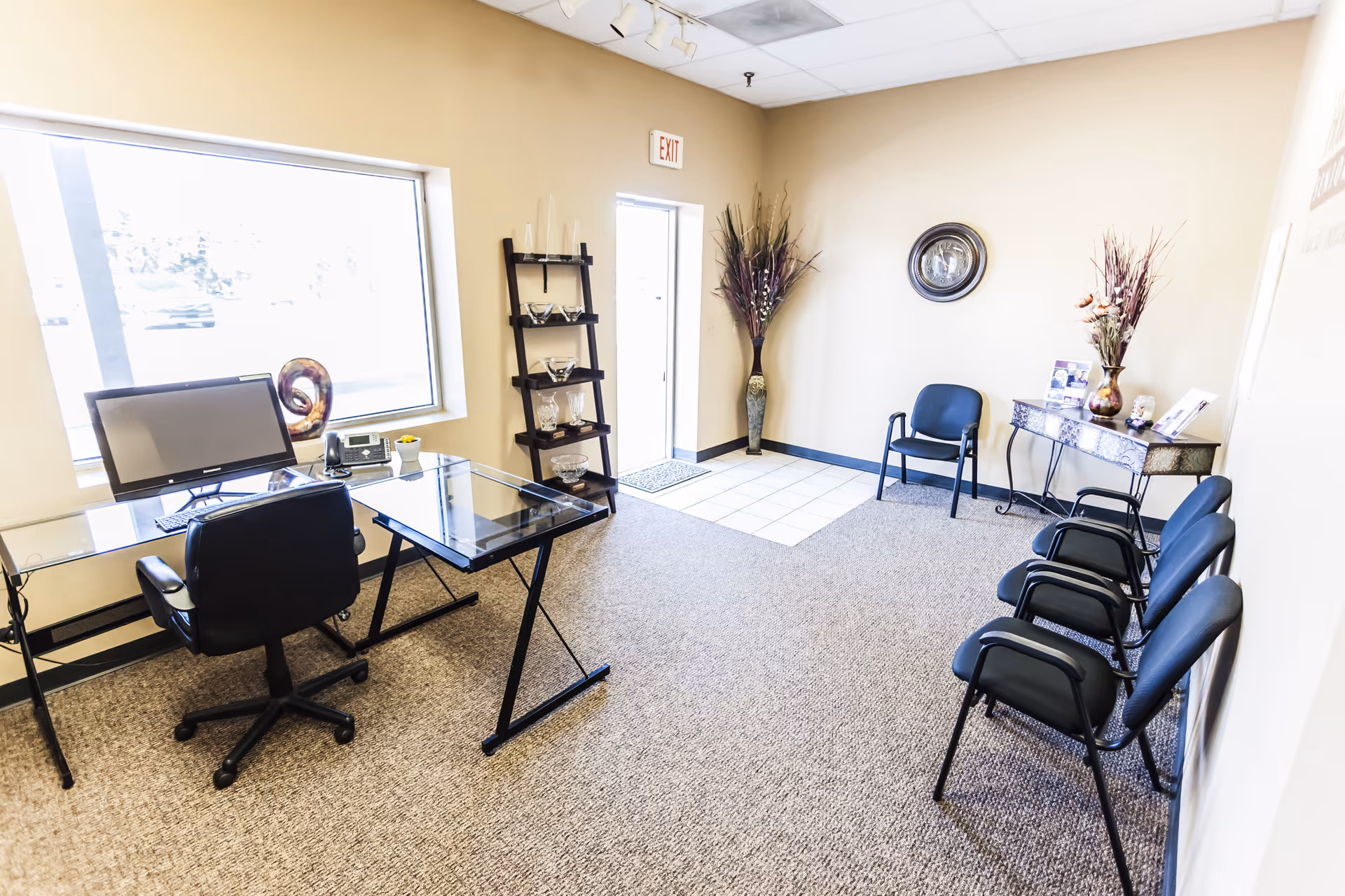 Reception area inside a facility featuring glass desk, computer, waiting chairs, and decorative plants.