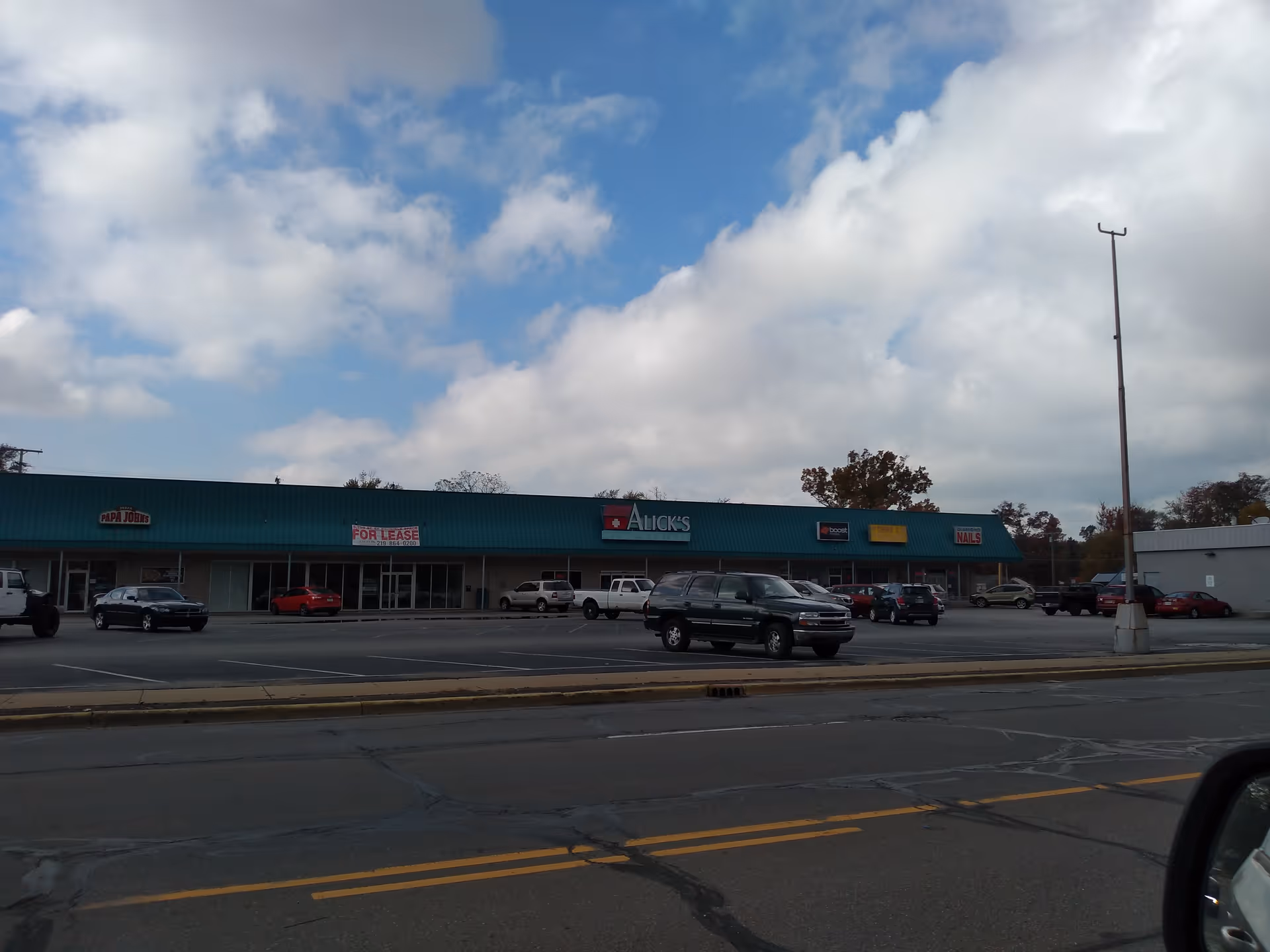Long strip mall storefronts with a teal roof, parking lot, and Alick's Home Medical sign.