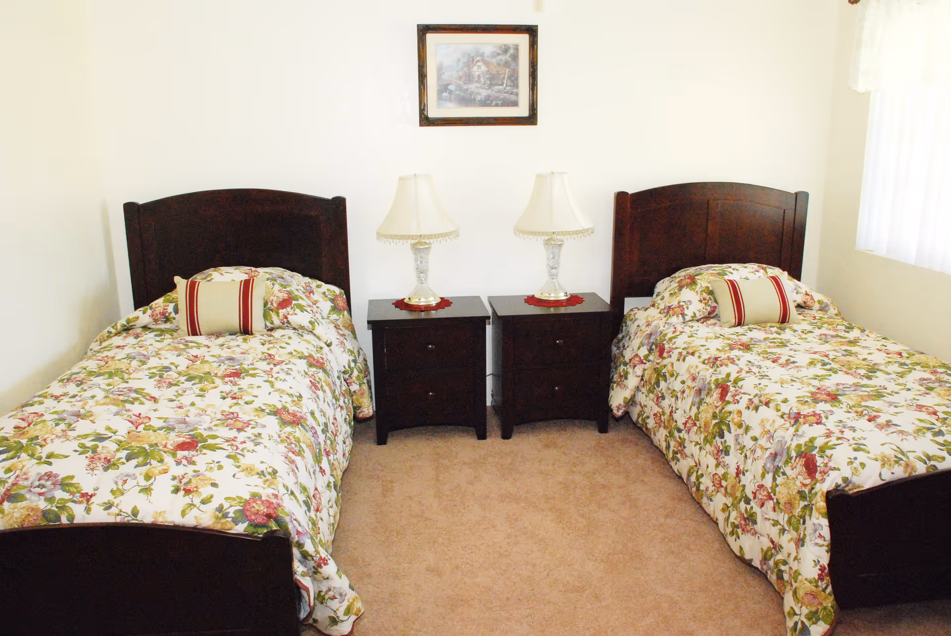A bedroom with two twin beds featuring dark wooden headboards and floral bedspreads. Between the beds are two matching dark wooden nightstands, each with a decorative lamp. A framed picture hangs on the wall above the nightstands. The room has light-colored walls and carpeted floor, with a window covered by white curtains on the right side.
