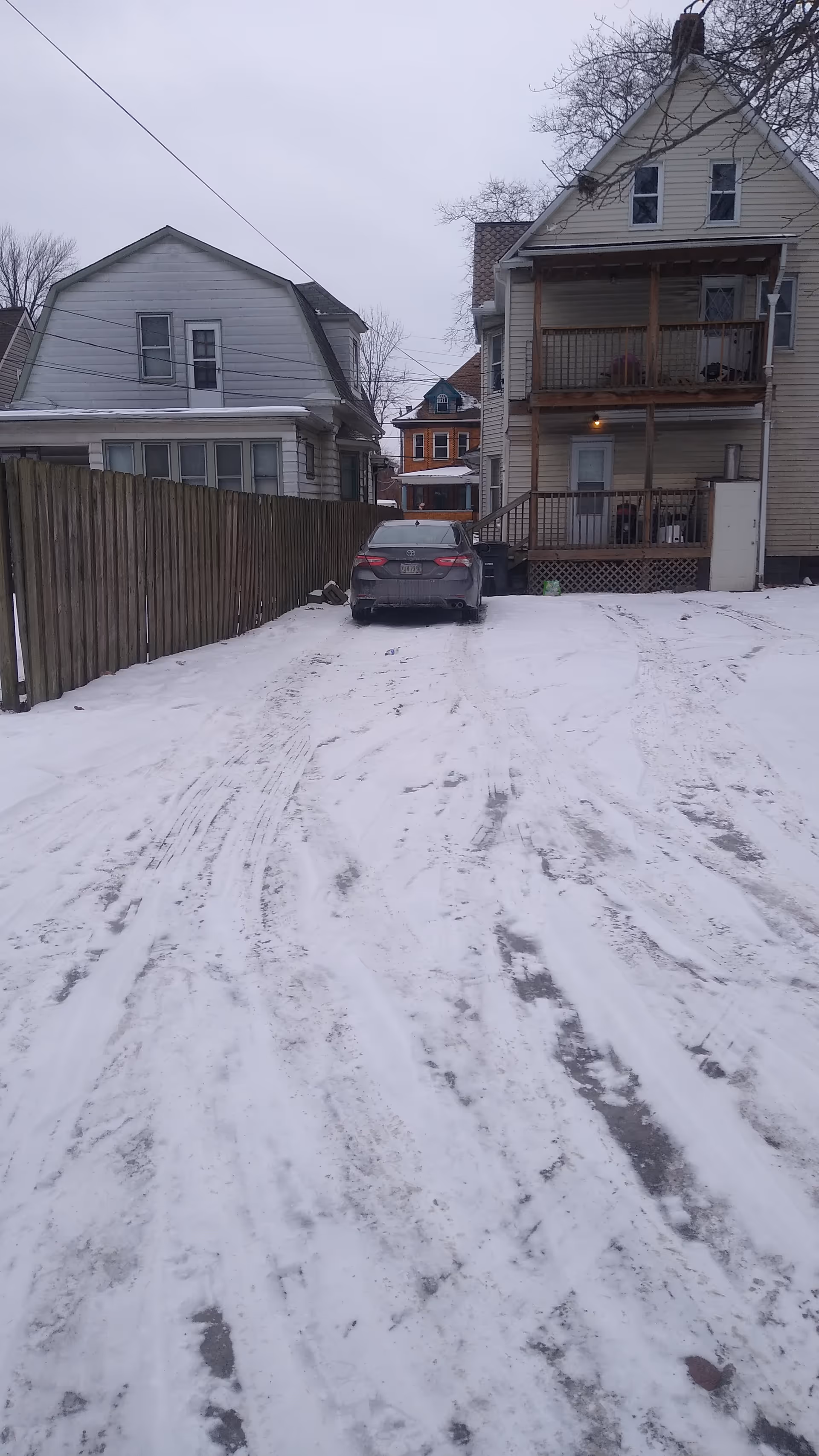 Snow-covered residential street with parked car and two wooden houses, one with a balcony.