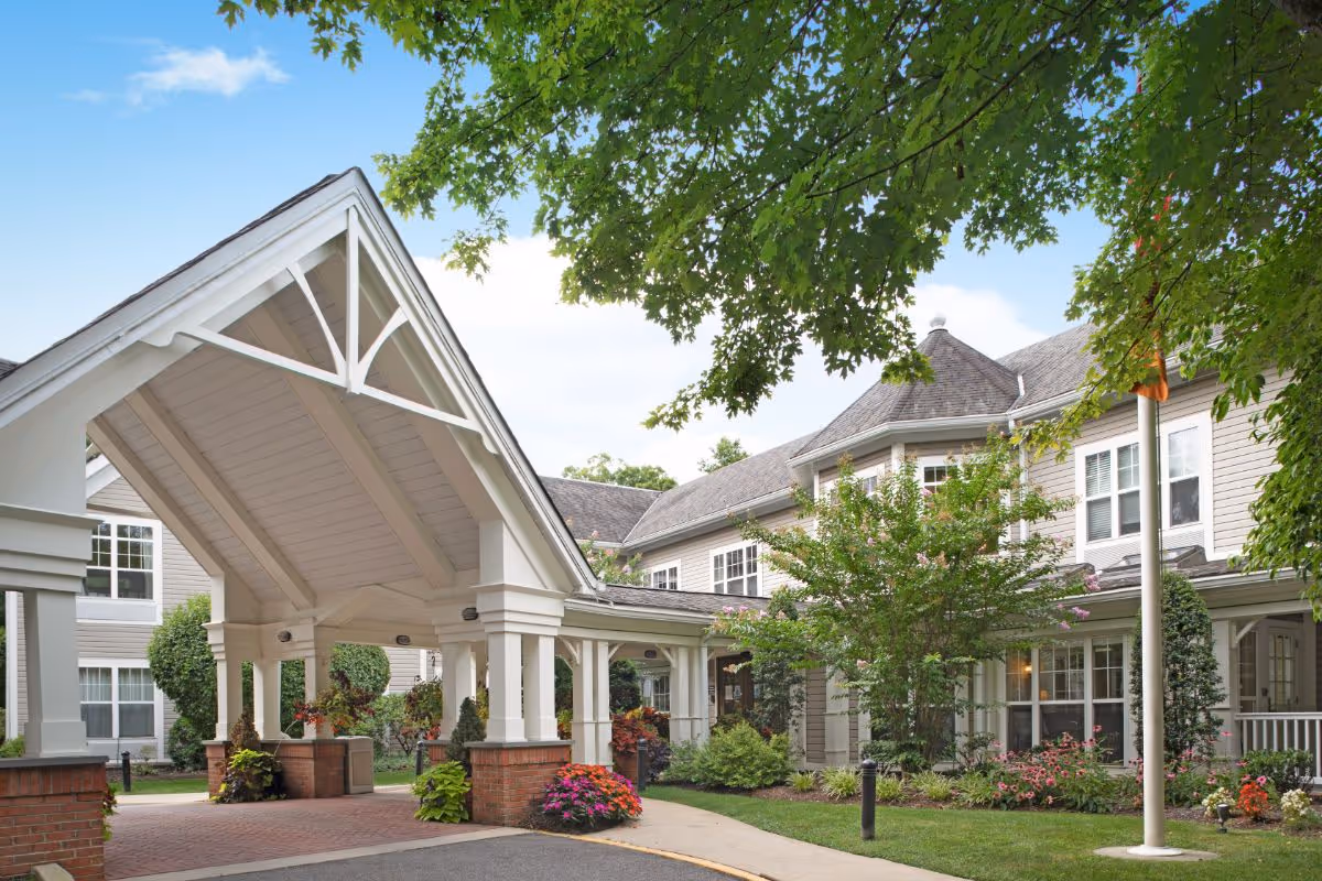 Entrance of a senior living facility with a covered driveway supported by white columns and brick bases, surrounded by well-maintained landscaping including green trees, shrubs, and colorful flowers under a clear blue sky.