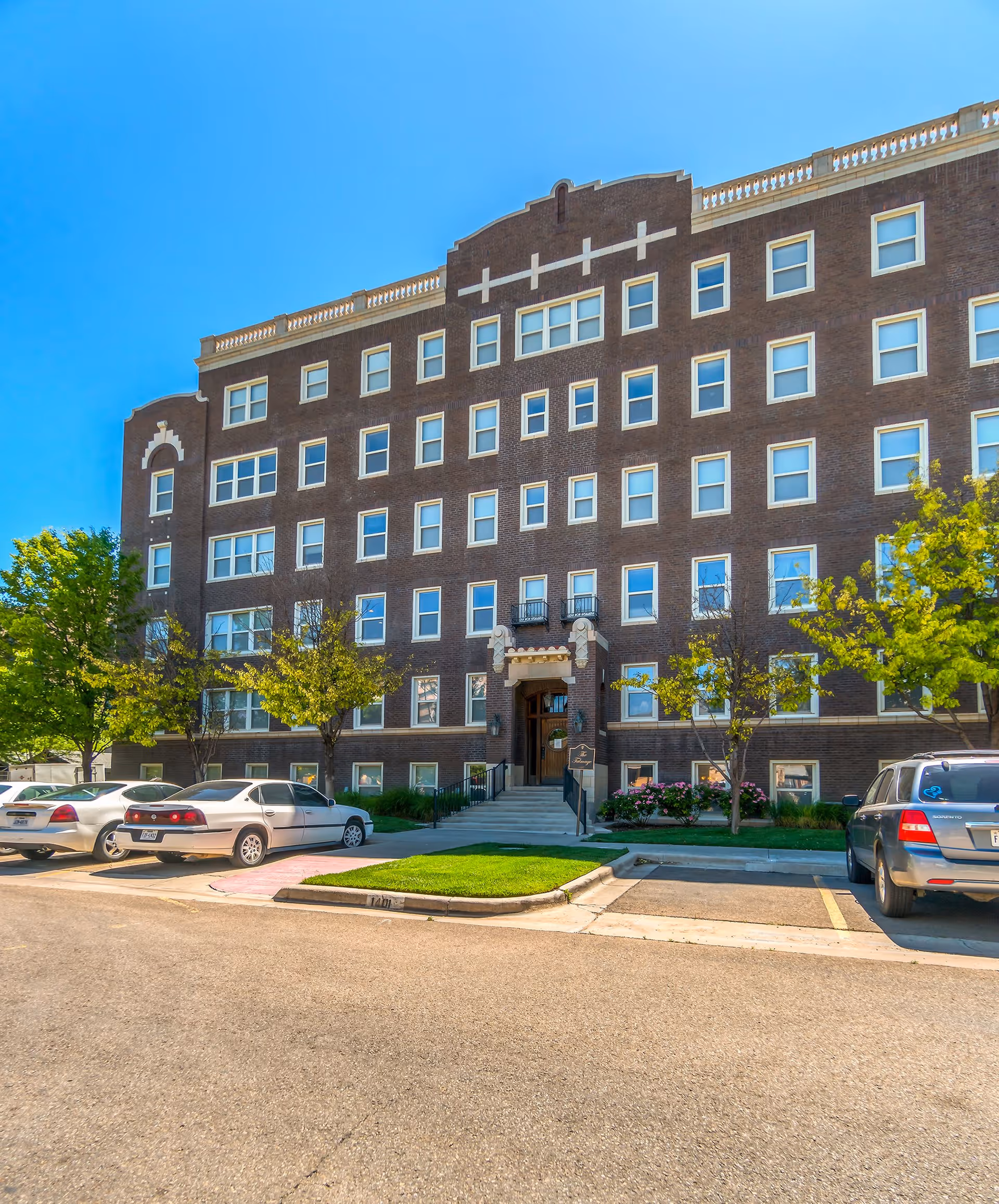 Exterior front view of a multi-story brick building with many windows, a central entrance with steps and railings, surrounded by small trees and parked cars under a clear blue sky.