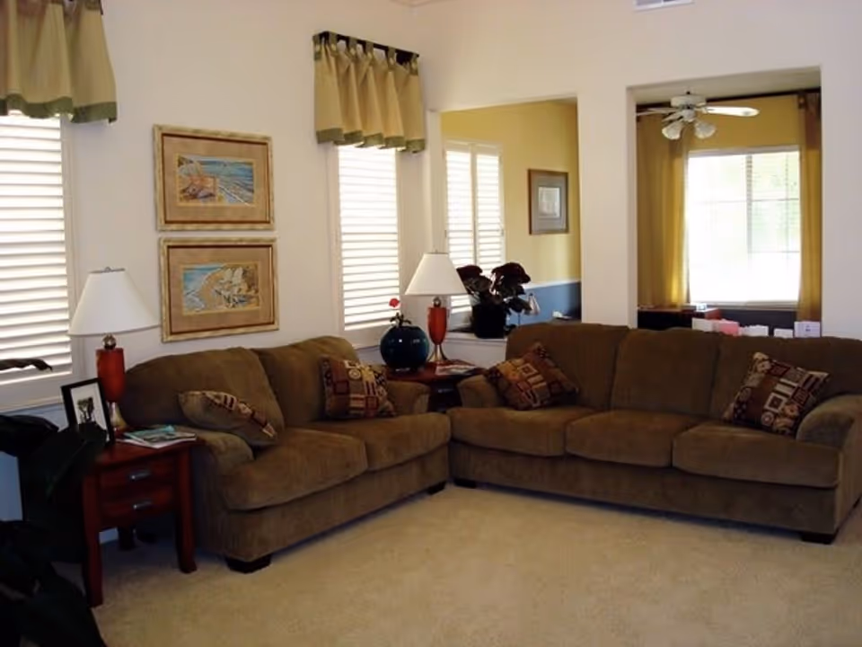 A cozy living room with two brown sofas arranged in an L-shape, each adorned with patterned throw pillows. There are two side tables with lamps, one with a red base and the other with a blue base. The walls are decorated with two framed paintings of beach scenes. The room has windows with white blinds and beige valances, and an adjoining room with yellow walls and a ceiling fan is visible through an open doorway.
