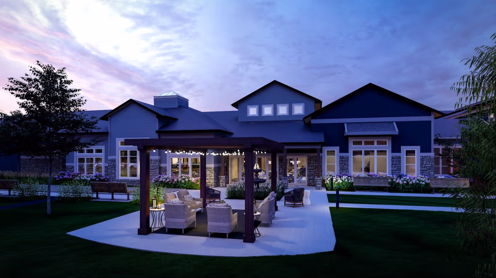 Dusk exterior of a senior living building with a lit pergola and outdoor seating on a landscaped lawn.