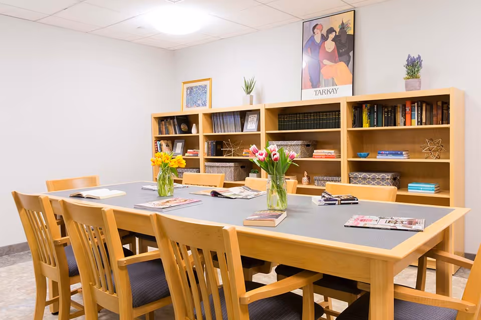 A bright communal room with a large wooden table surrounded by chairs, vases of flowers, and bookshelves along the back wall.