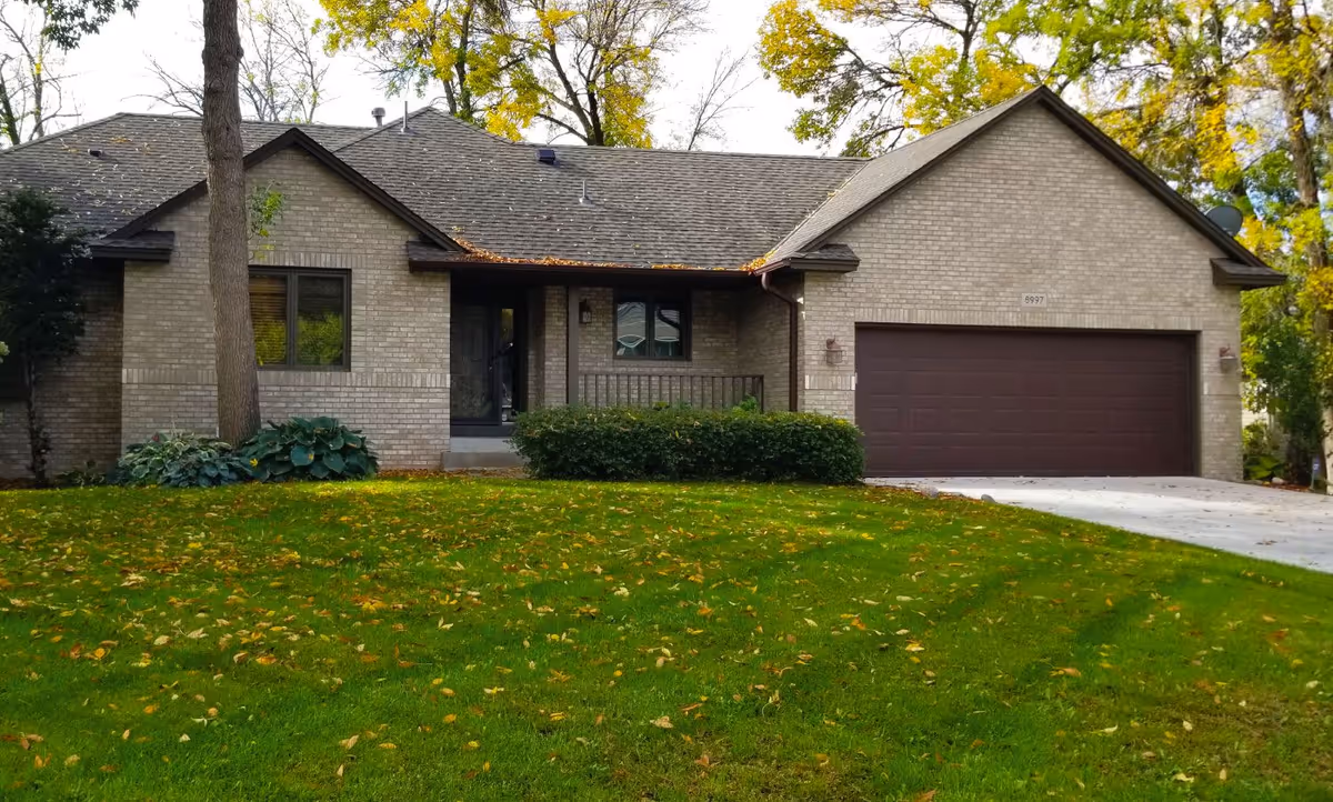 Exterior front view of a single-story brick house with a two-car garage, a green lawn with scattered fallen leaves, and trees in the background.