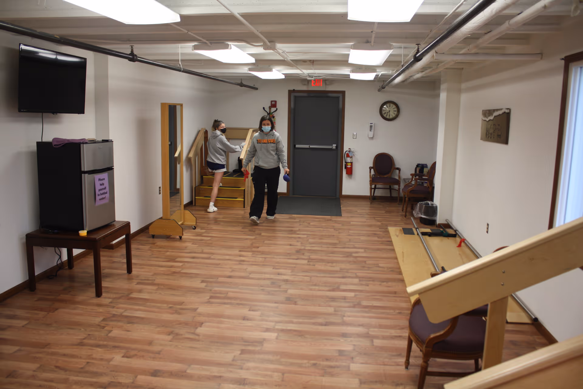 An open interior activity room with wood floors showing a small TV and fridge, chairs and exercise equipment, and two masked people near a doorway.