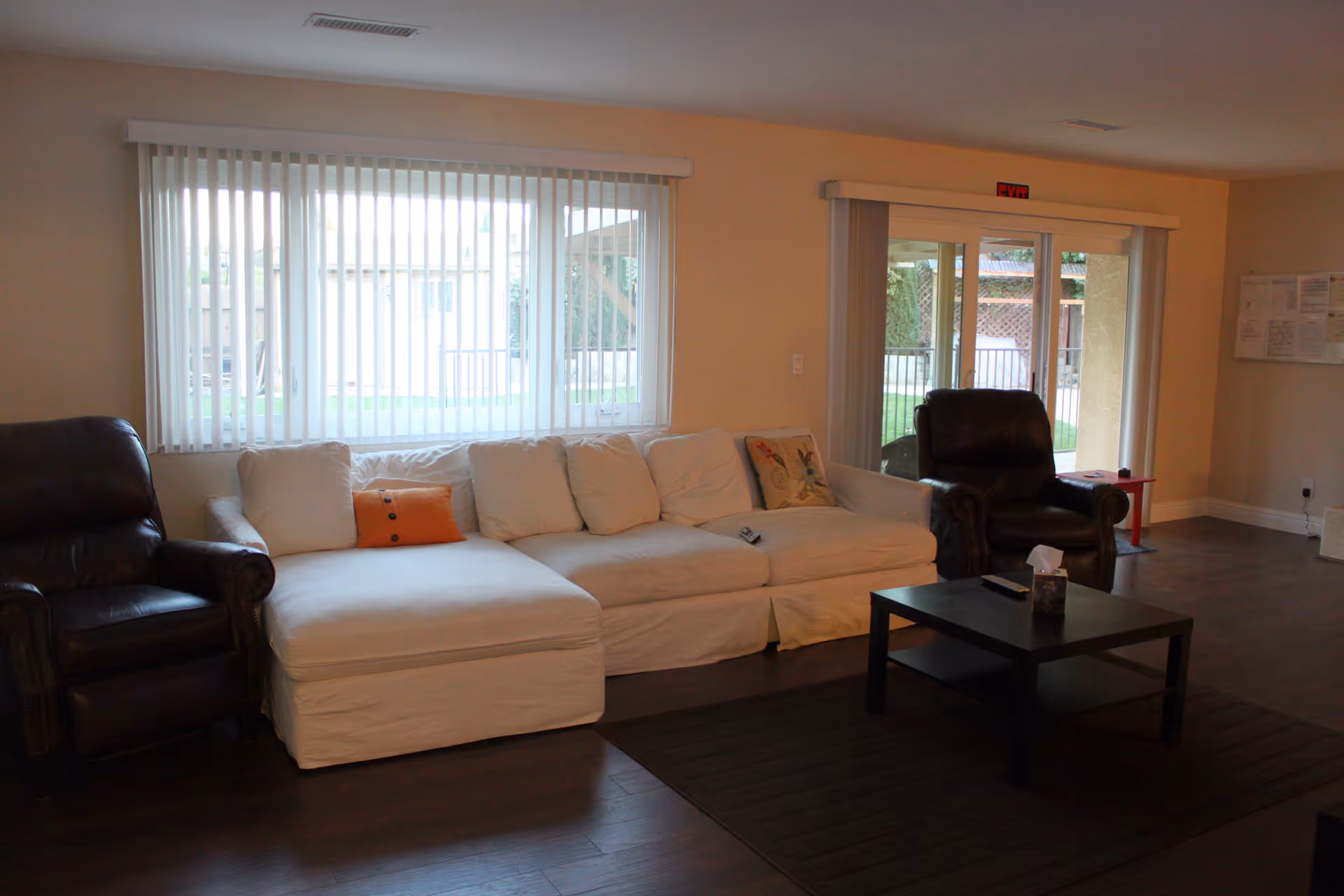 A living room with a white sectional sofa, two dark leather recliners, a coffee table, and large windows with vertical blinds.