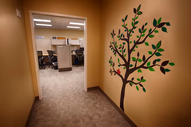 Beige hallway featuring a tree mural leads to a cubicle-filled office area.