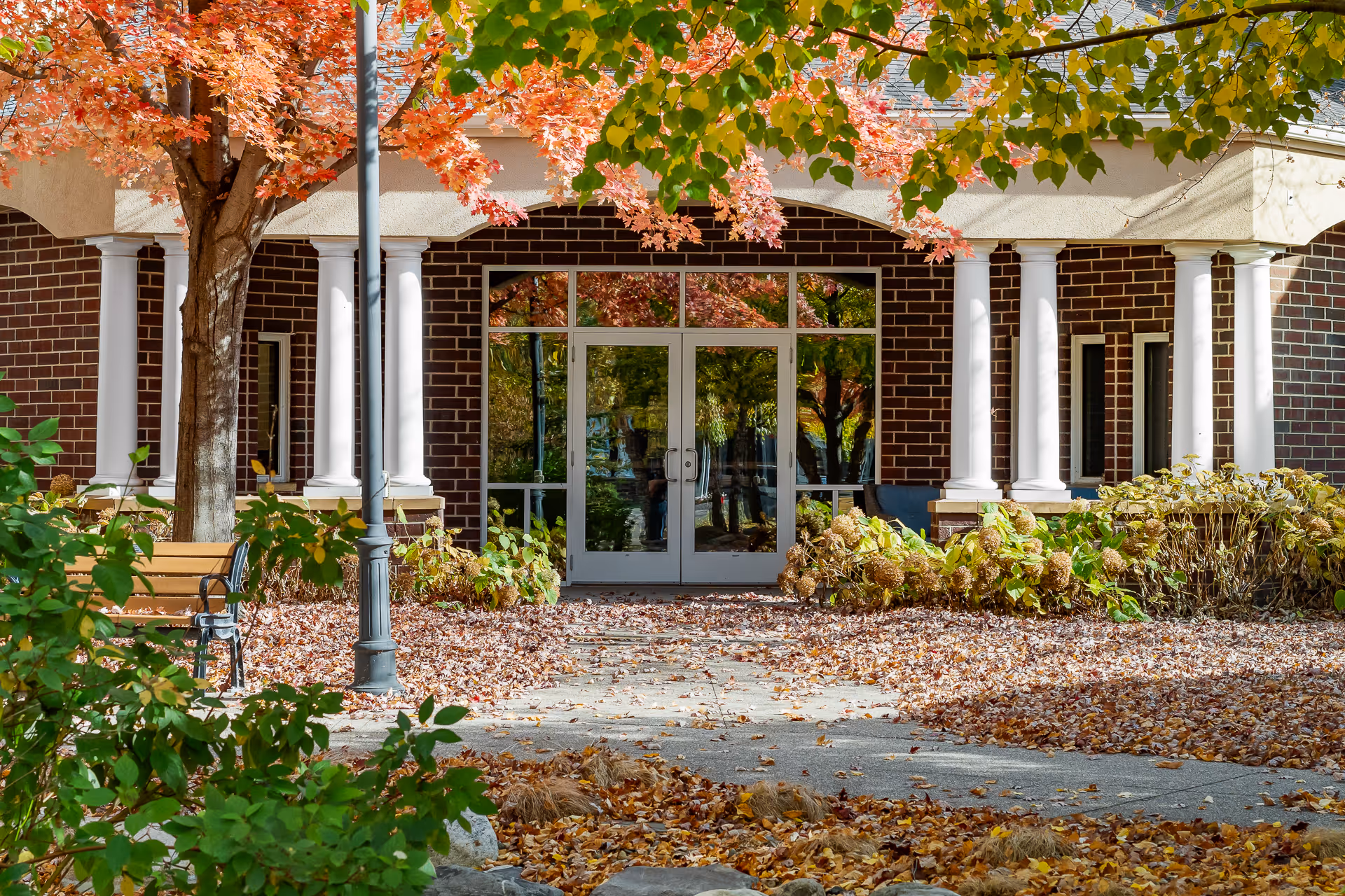 Entrance to a building with glass double doors framed by white columns and brick walls, surrounded by autumn foliage with fallen leaves on the ground and a bench to the left.