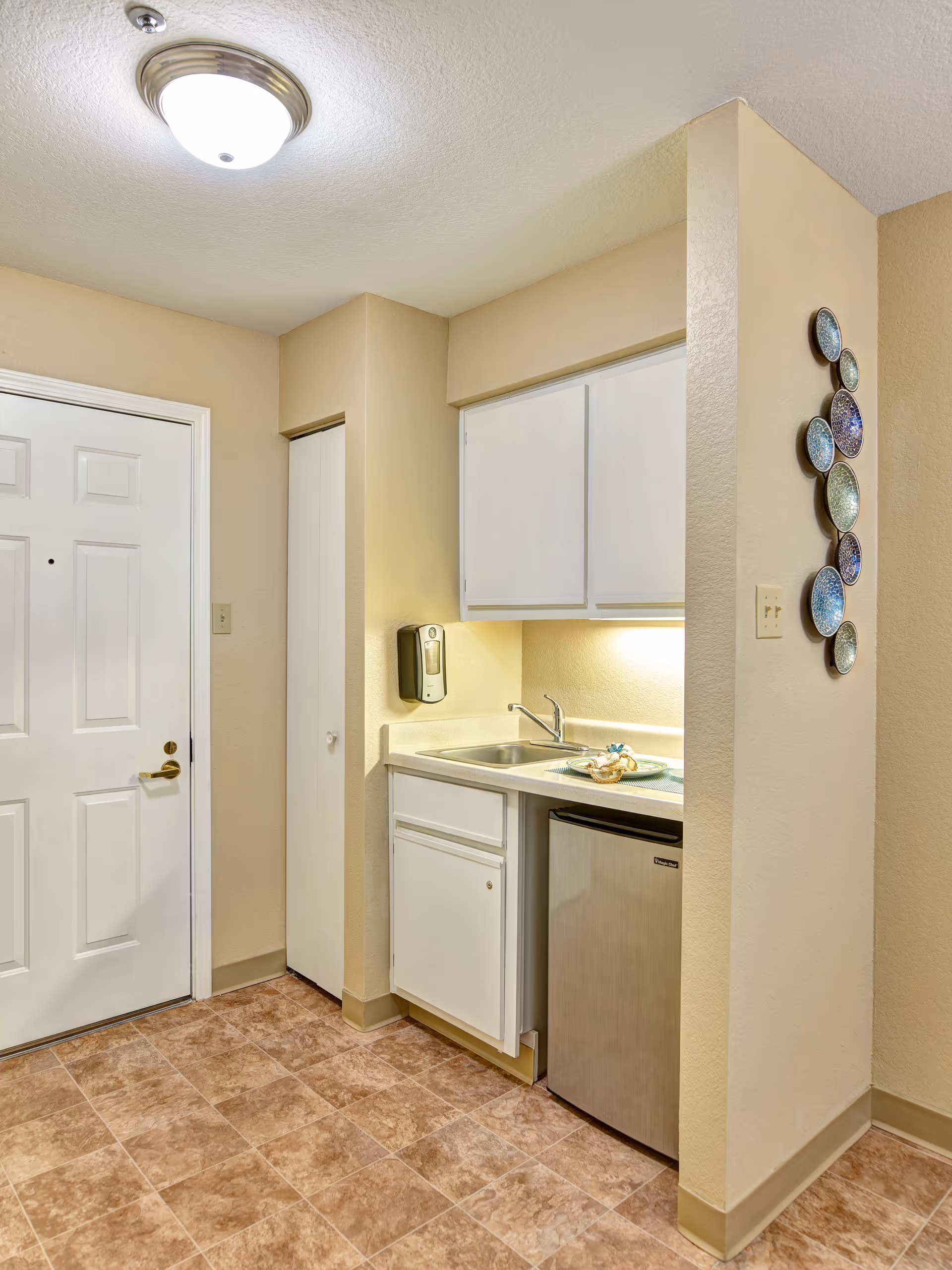 Small kitchenette area with a white door on the left, a small sink with a faucet, white cabinets above and below the counter, a small stainless steel refrigerator, and a wall-mounted soap dispenser. The floor has brown tile, and there is decorative wall art with circular plates on the right wall.