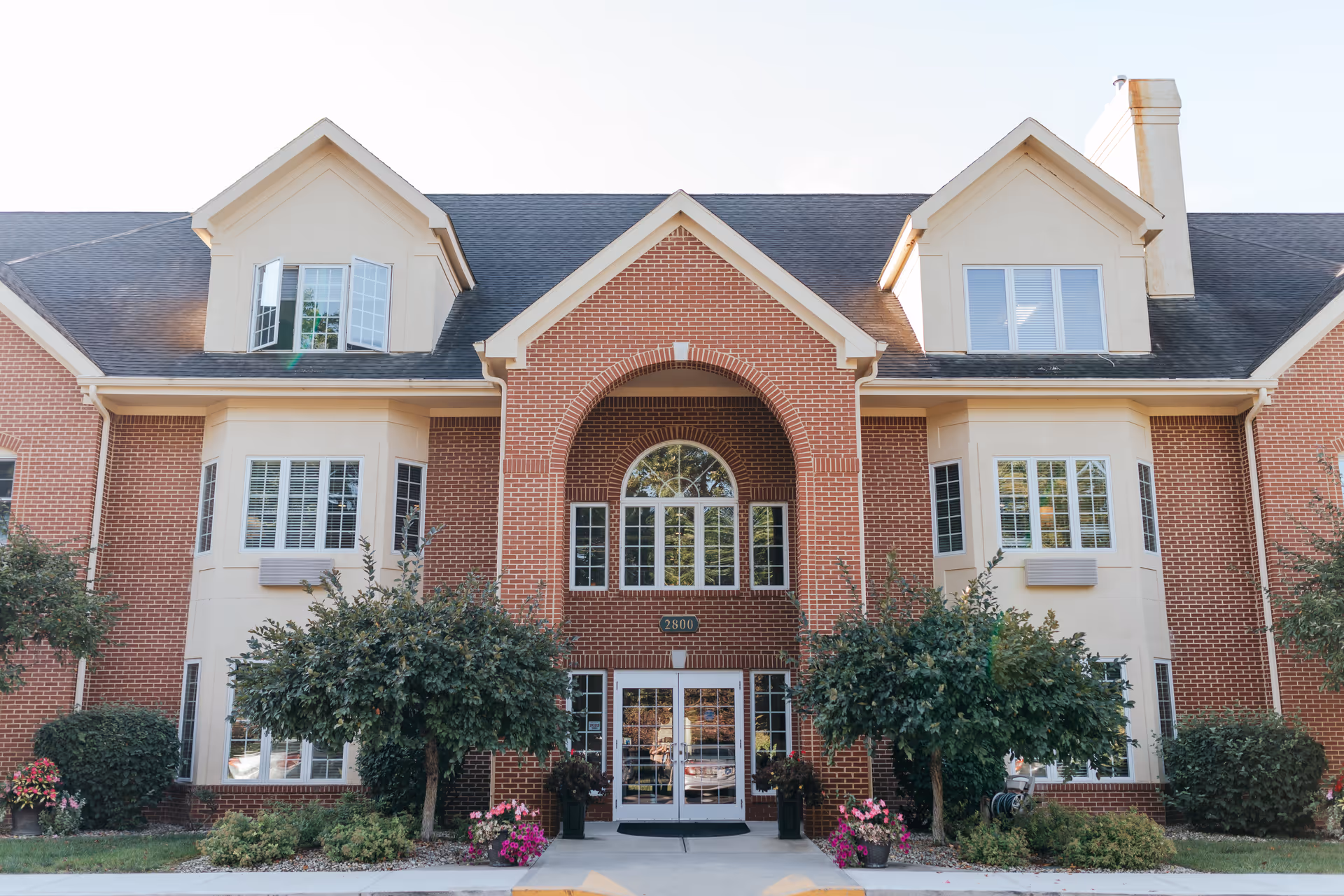 Front exterior view of a senior living facility named Senior Living at Forest Ridge, featuring a brick building with large windows, a central arched entrance, and landscaping with bushes and flowers.
