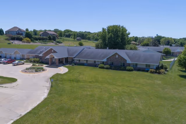 A single-story senior living facility building with a stone and siding exterior, surrounded by a large green lawn and a circular driveway with parked cars. Trees and other houses are visible in the background under a clear blue sky.