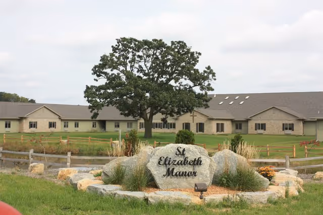 Exterior of a long, single-story senior living facility with a large tree and stone sign.