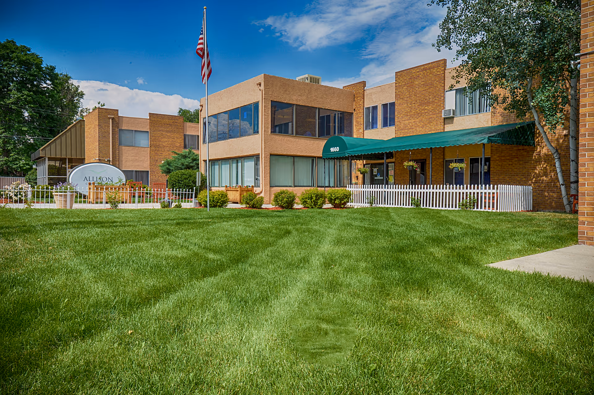 Exterior view of Allison Care Center, a two-story brick building with large windows, a green awning over the entrance, a white picket fence, a well-maintained green lawn, and an American flag on a flagpole in front.