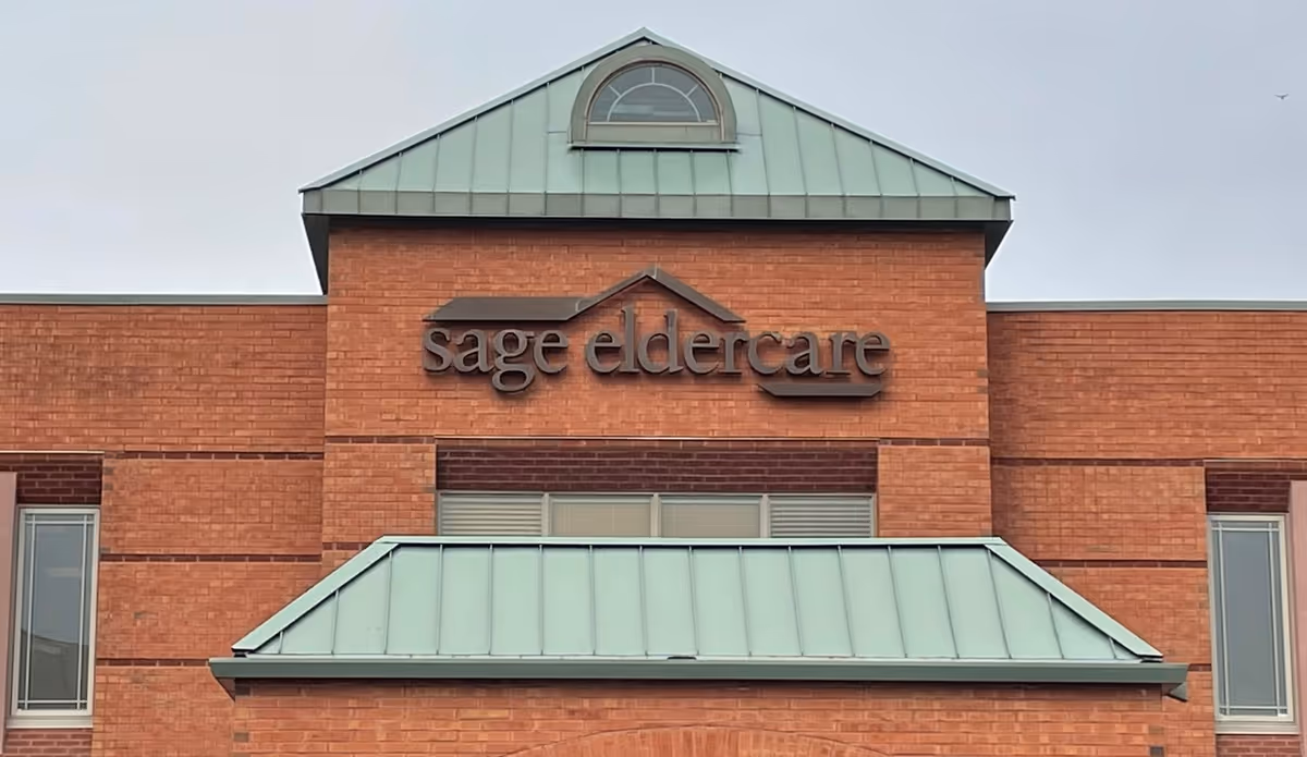 Brick Sage Eldercare building front with teal metal roof and sign.