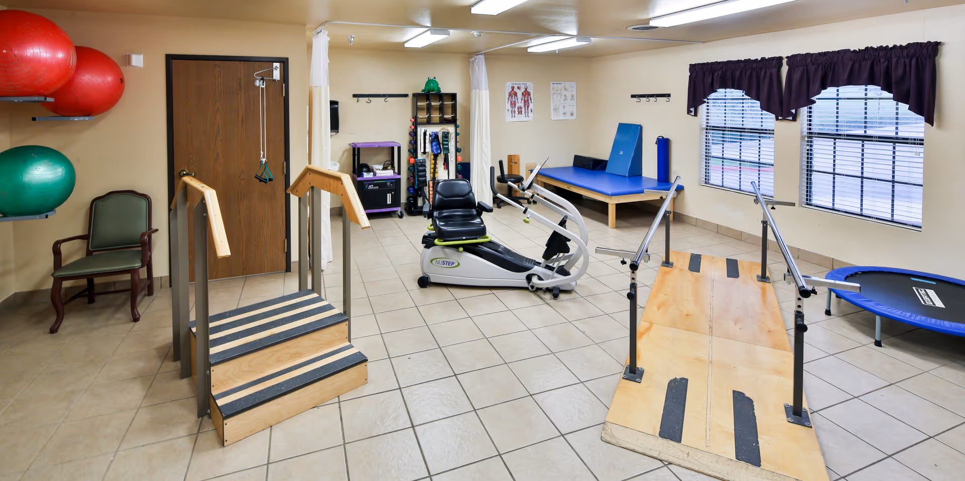A physical therapy room with exercise equipment including a small wooden staircase with handrails, a recumbent exercise bike, a wooden ramp with handrails, a trampoline, and large exercise balls mounted on the wall. There is a chair in the corner, anatomical charts on the wall, and windows with dark curtains letting in natural light.