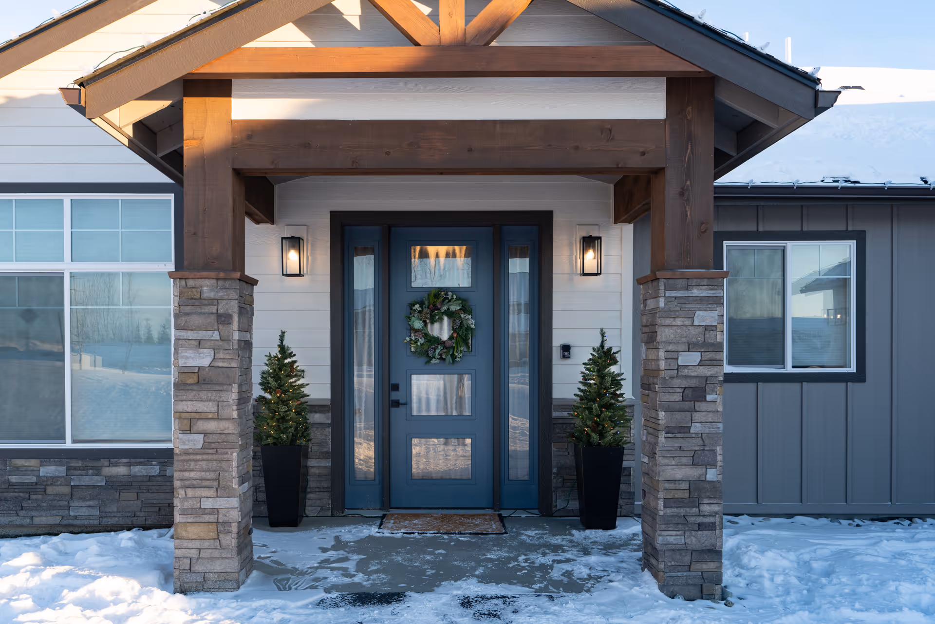 Front entrance of a building with a blue door decorated with a green wreath. The entrance is framed by wooden beams and stone pillars, with two potted evergreen plants on either side. Snow is visible on the ground and roof.