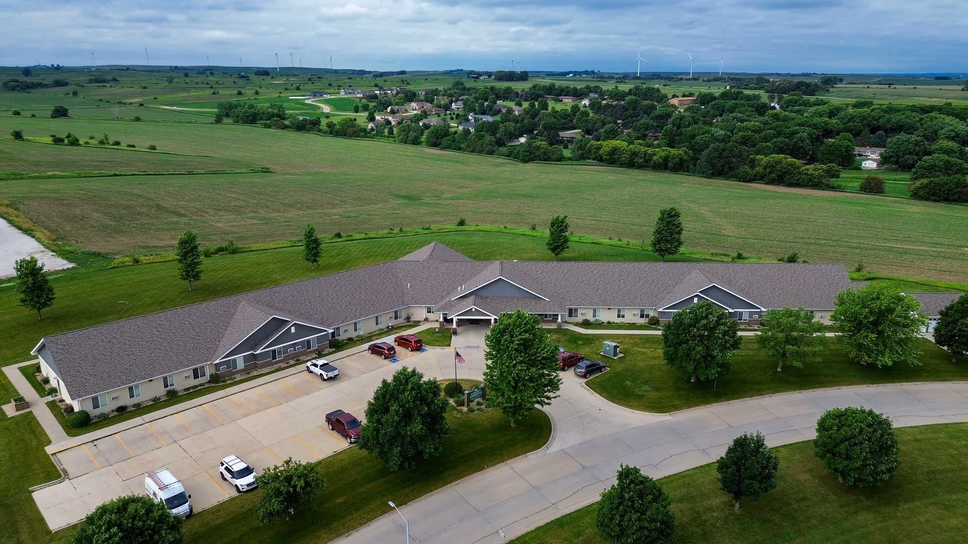 Aerial view of a single-story senior living facility named Golden Horizons of Ida Grove surrounded by green lawns and trees, with a parking lot in front and expansive farmland in the background under a cloudy sky.