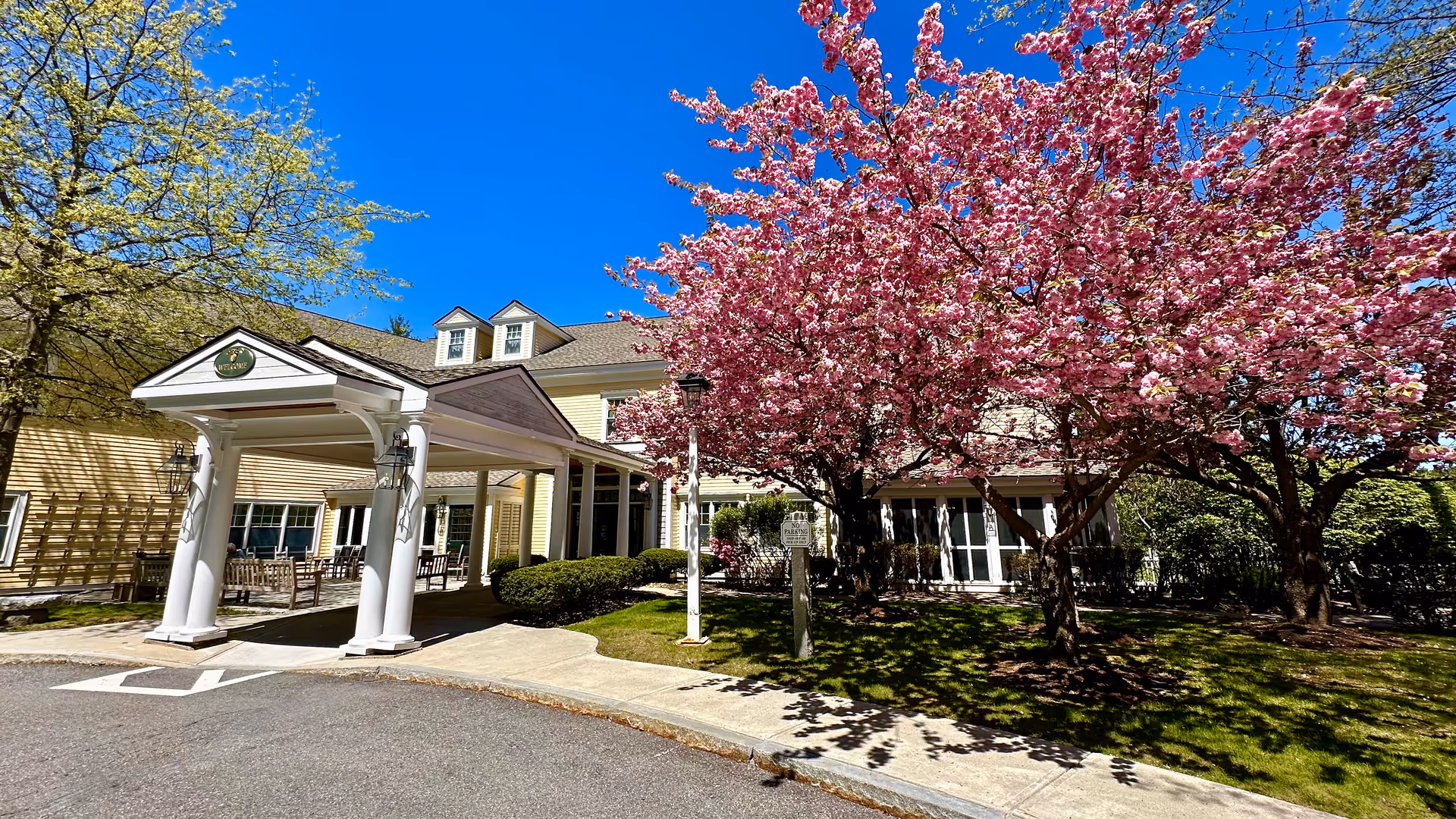 Exterior view of Orchard Hill Sudbury Assisted Living with pink blossoms and covered entrance.
