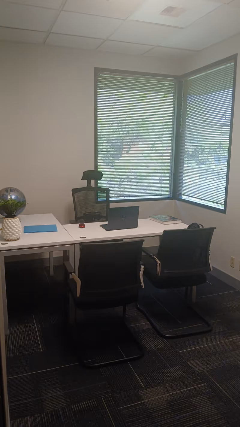 A small office corner with white desks, two chairs, laptop, plant, and large window blinds.