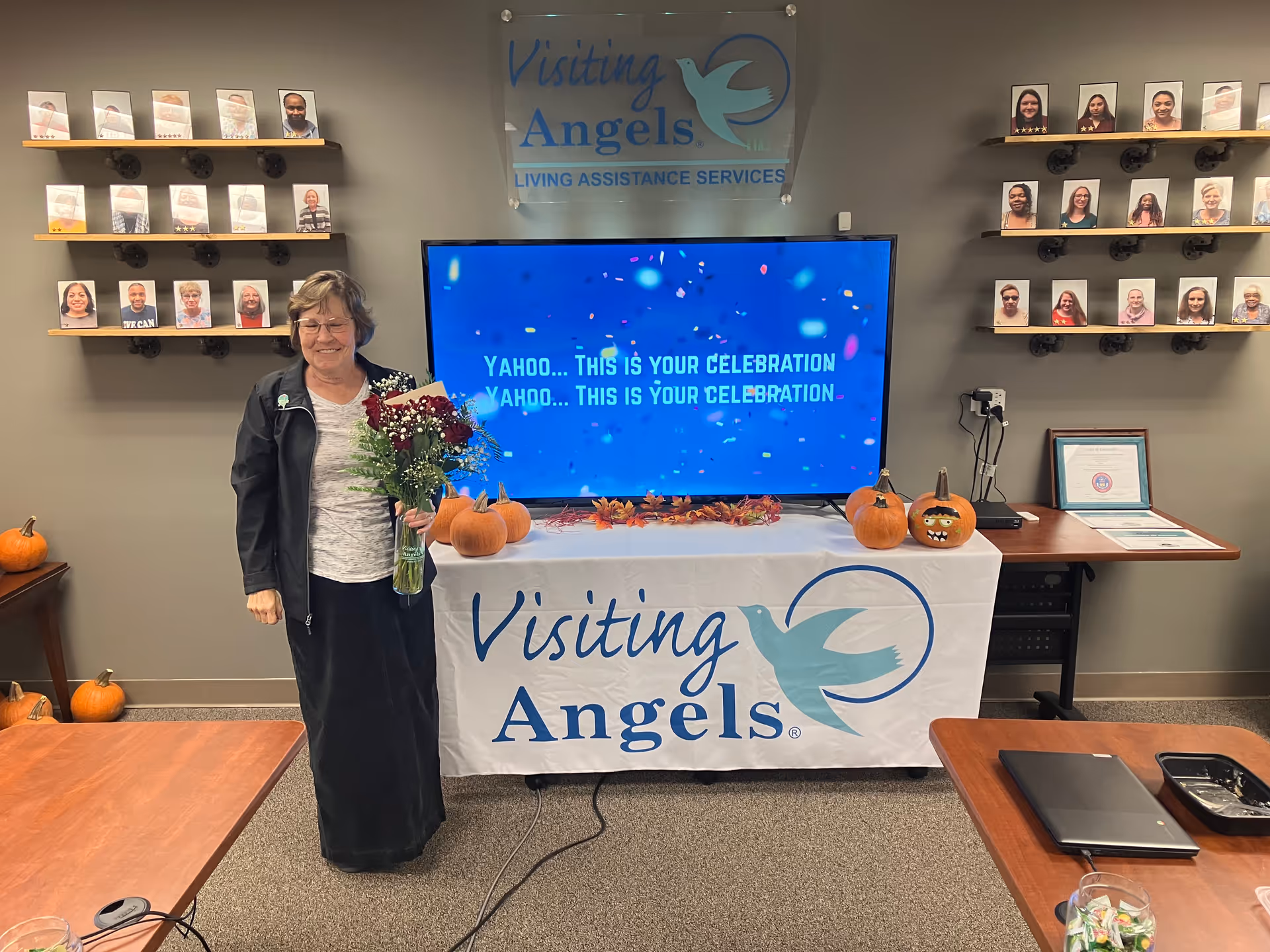Smiling staff member stands beside a celebration setup and TV inside Visiting Angels lobby.