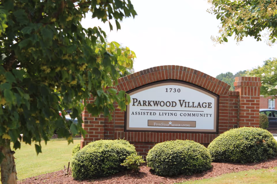 A brick sign at the entrance of Parkwood Village Assisted Living Community with neatly trimmed bushes and trees around it under a clear sky.