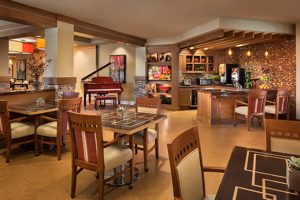 A warm and inviting common area in a senior living facility featuring several wooden tables and chairs with cushioned seats. In the background, there is a red grand piano, a kitchen area with a mosaic tile backsplash, countertop appliances, and a beverage dispenser. Decorative shelves with vases and plants add to the cozy atmosphere.