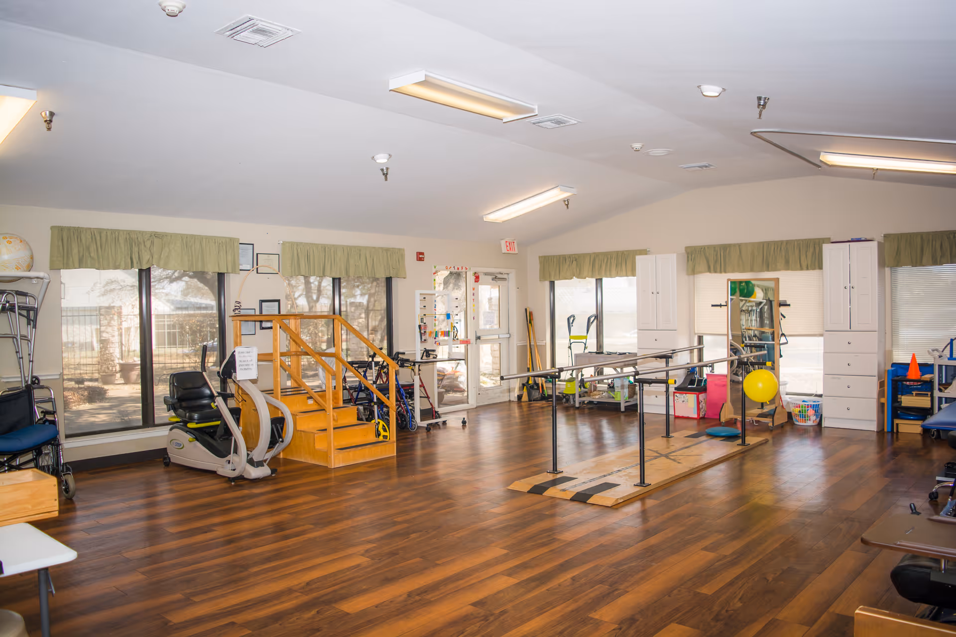 Spacious therapy room with wooden floors, exercise equipment, parallel bars and large windows.
