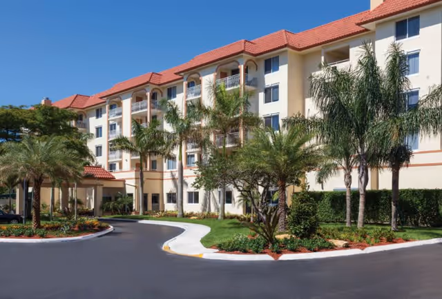 Exterior front of a multi-story senior living building with a red tile roof, balconies, palm trees, and a circular driveway.