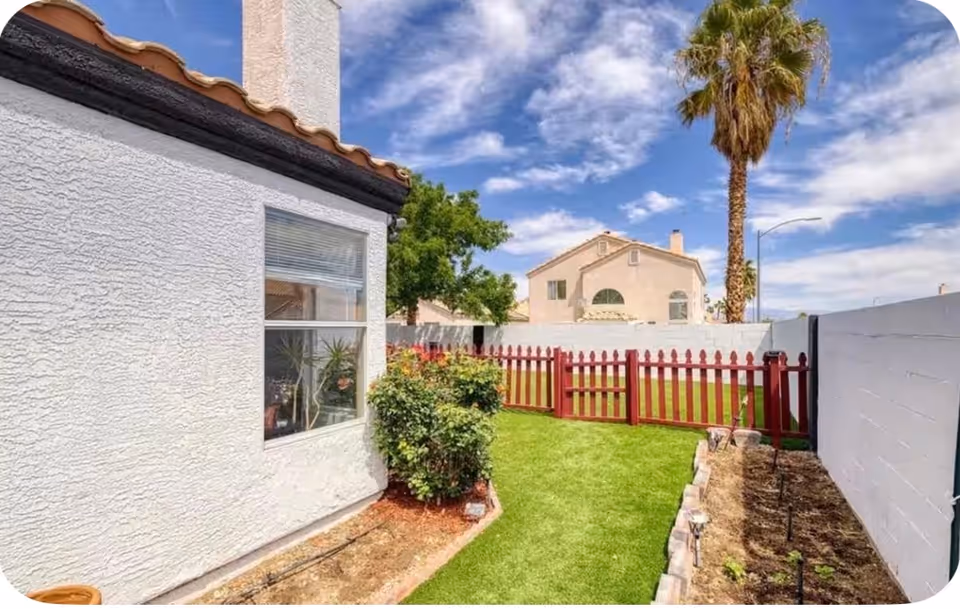 White stucco house with green lawn, red fence, and palm tree in a sunny backyard.