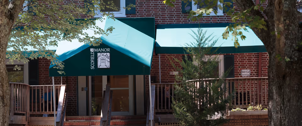 Entrance of Scofield Manor with green awnings displaying the facility name and logo, surrounded by trees and a wooden railing.
