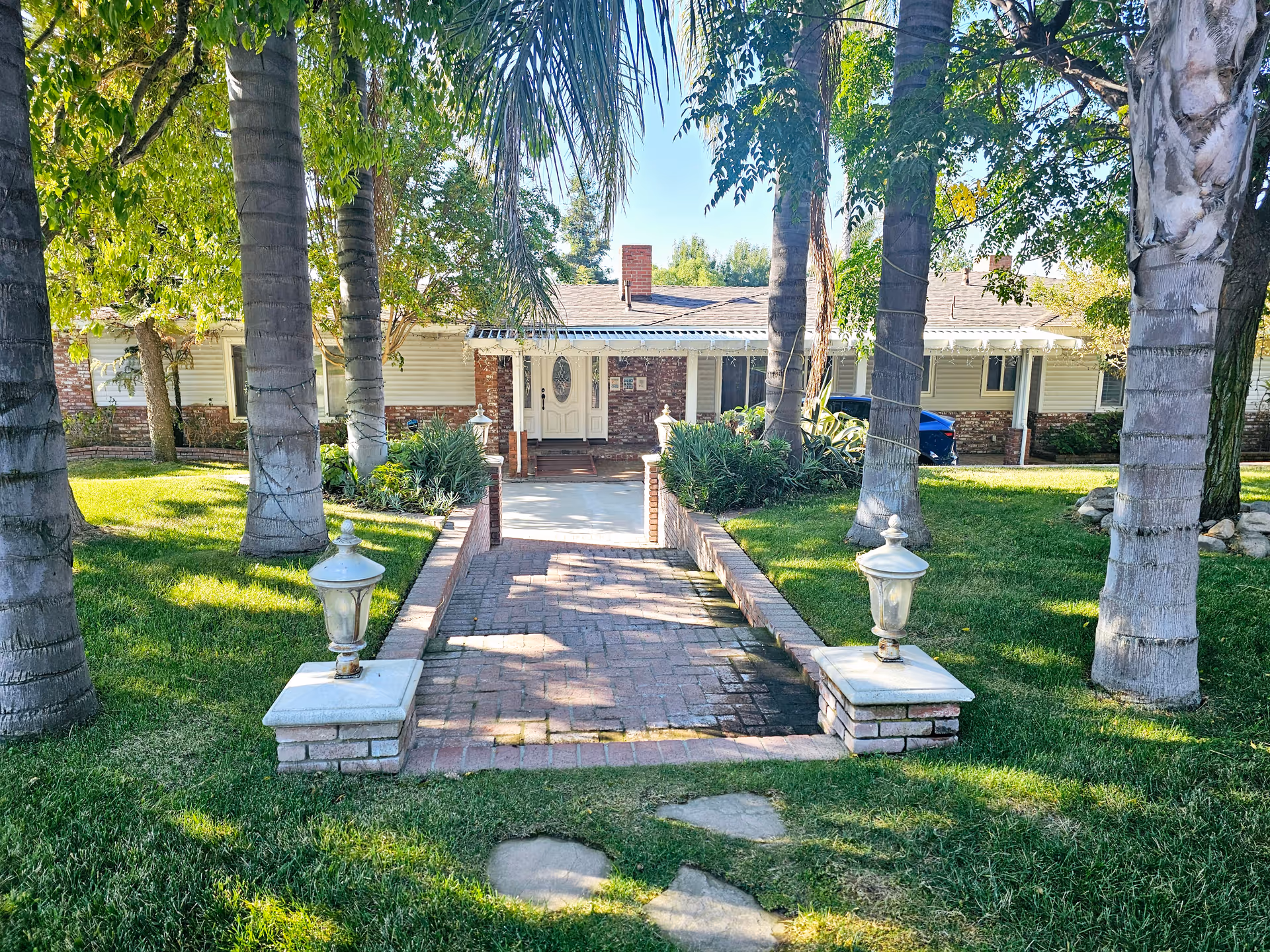 Front exterior view of a single-story building with a brick pathway leading to a white door entrance. The pathway is flanked by two decorative lanterns on brick pedestals. The area is surrounded by green grass, palm trees, and other plants under a clear blue sky.