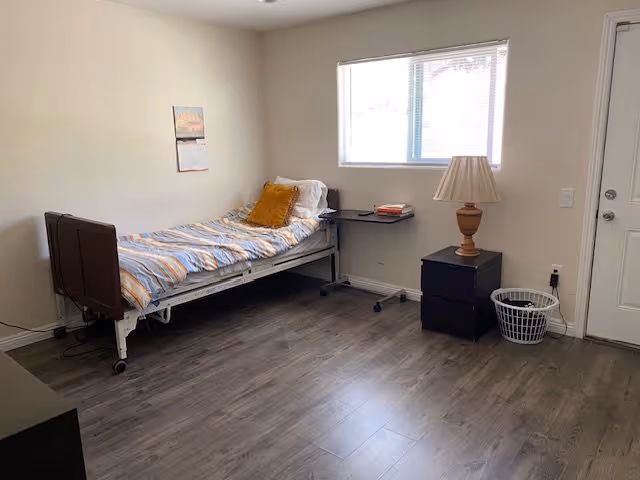 Simple furnished bedroom with a single bed, bedside table and lamp, window, and wood-look flooring.