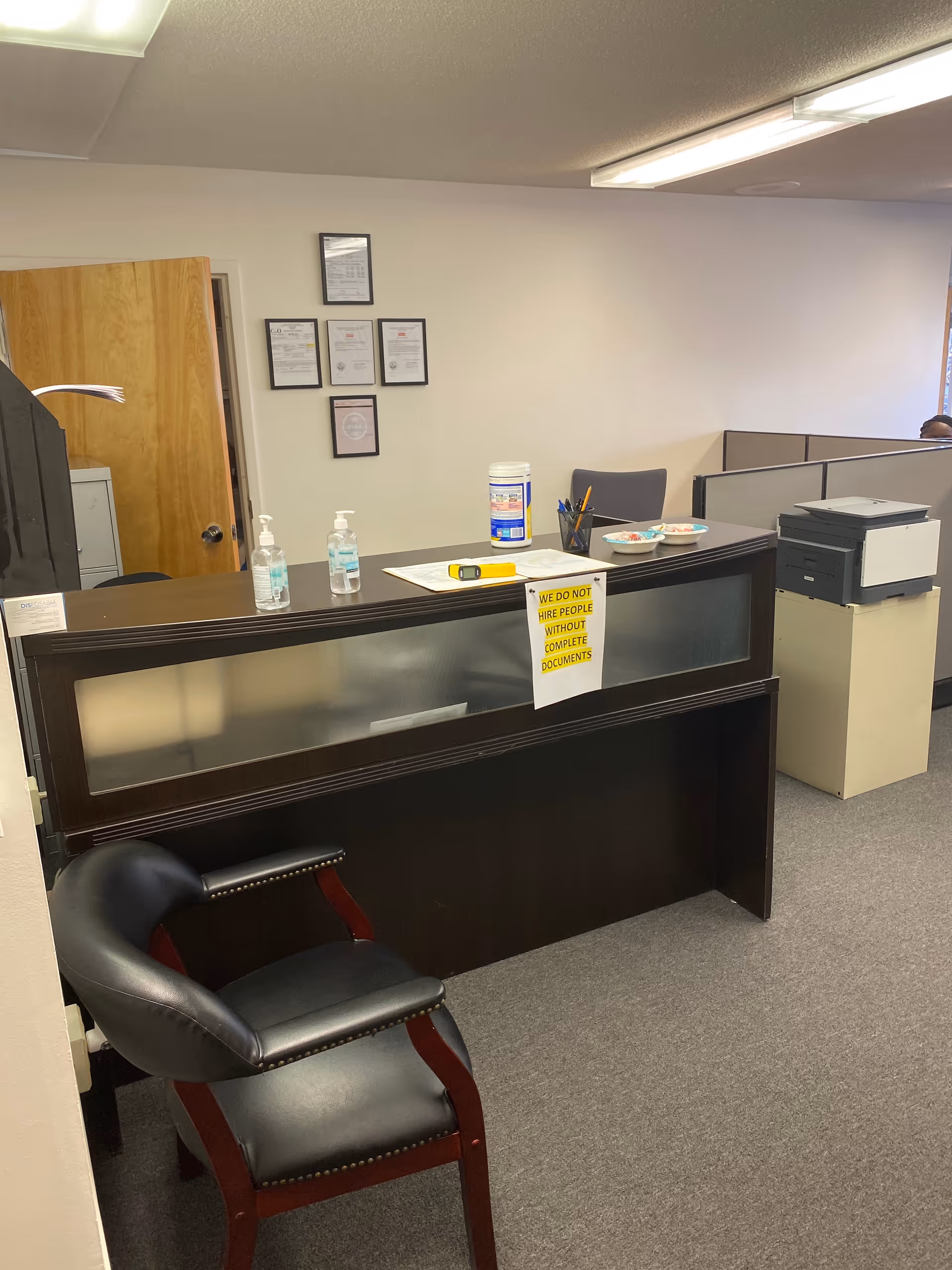Reception desk with frosted glass panels, a black chair, and framed certificates on a pale wall.