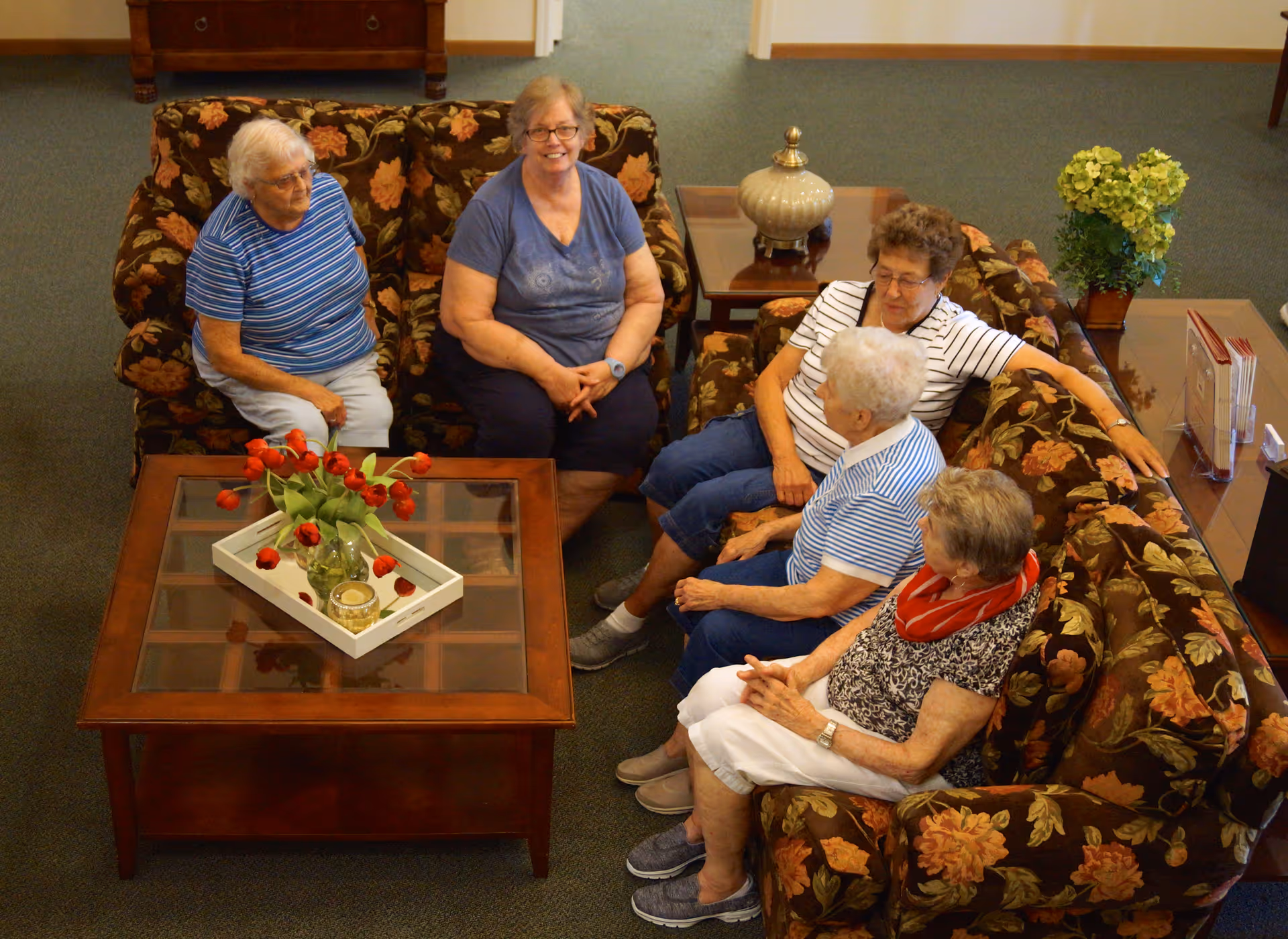 Five elderly women sitting and chatting on floral patterned couches around a wooden coffee table with a glass top, which has a vase of red tulips and a candle on a white tray. The room has green carpet and wooden furniture, including a side table with a lamp and a small plant.