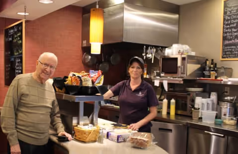 A staff member and a resident standing at a service counter in a kitchen/dining area with snacks and cooking equipment.