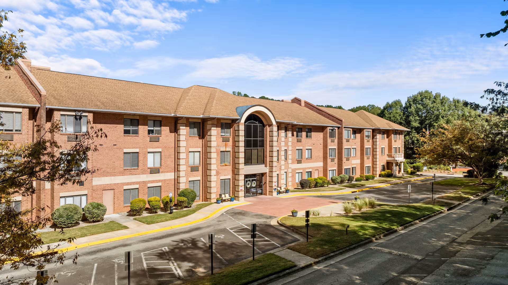 Sunlit brick senior living building exterior with a central entrance and driveway.