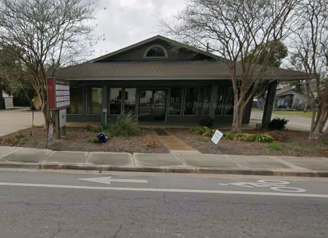 Single-story building front with glass doors, surrounding landscaping, and a street in view.