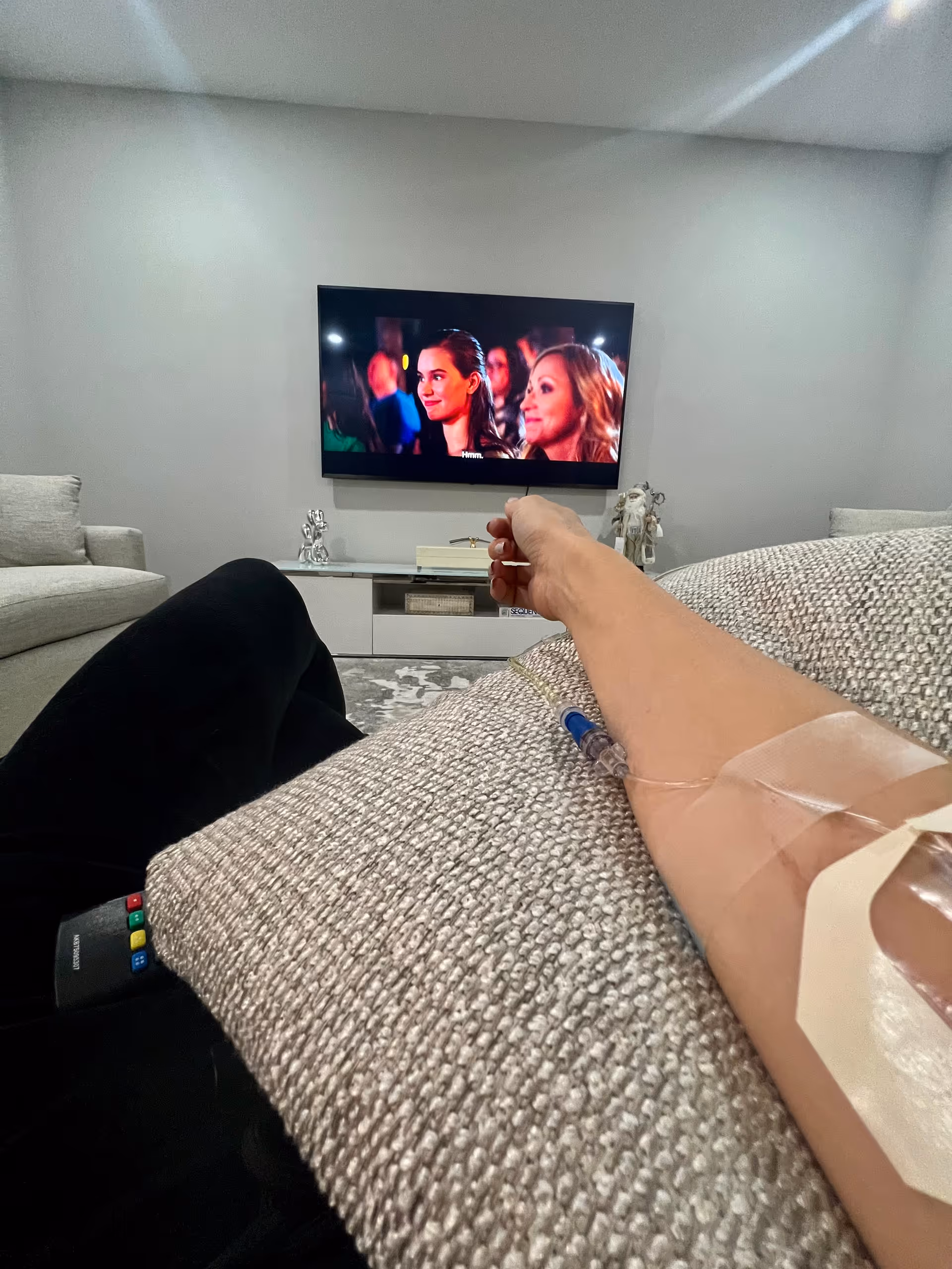 Person with IV line reclines on a couch watching TV in a modern living room.