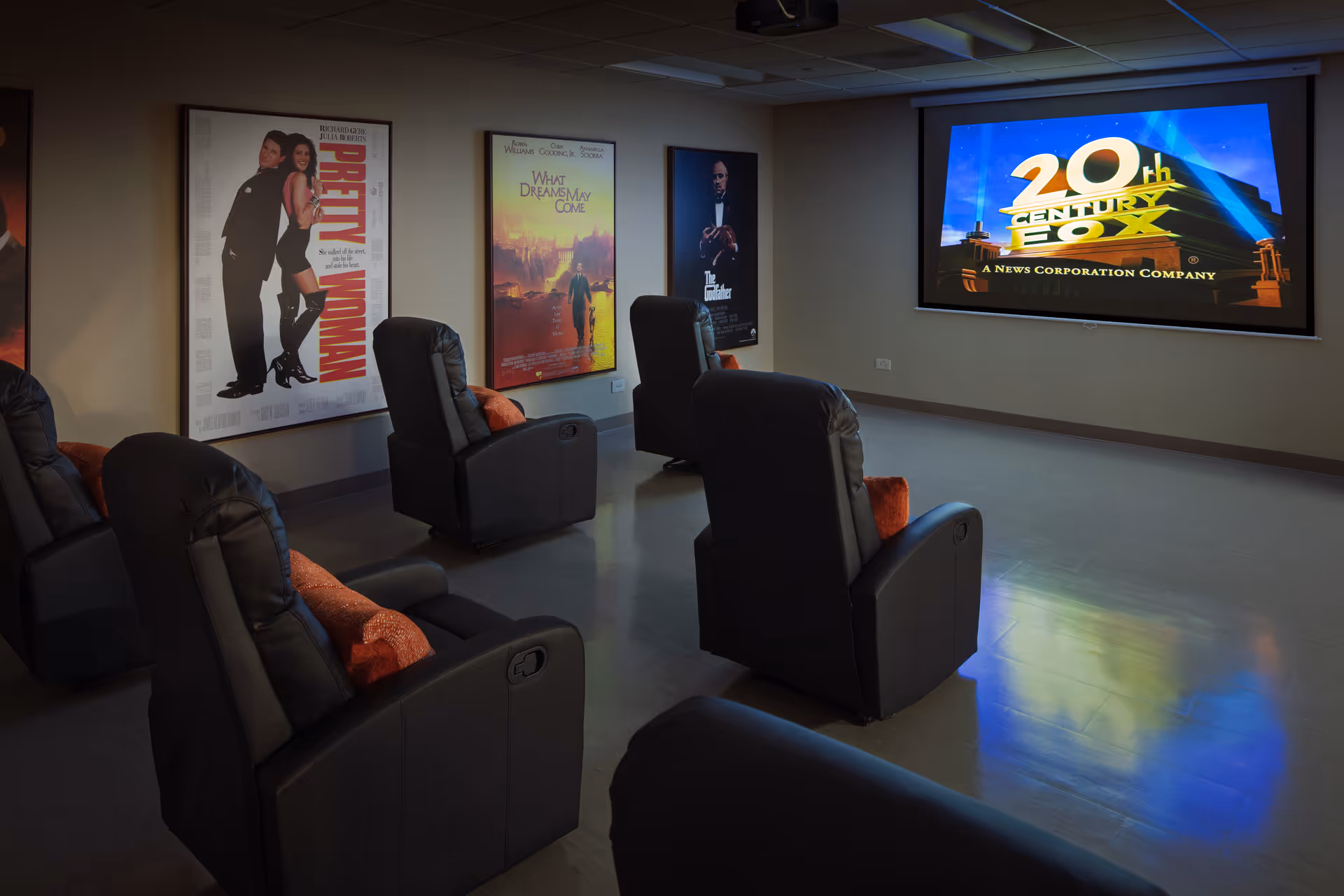 Dimly lit senior living center theater room with recliners, posters, and a large screen.